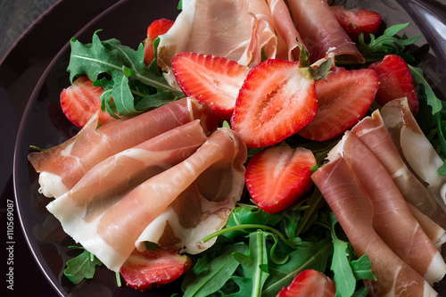 Fresh salad with strawberry, arugula and prosciutto on purple plate. Low key