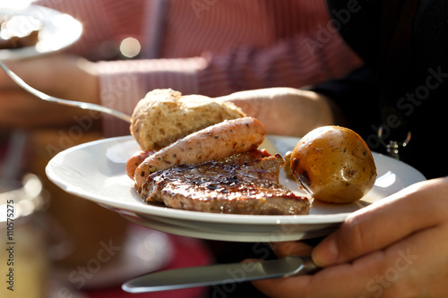Hand holding a plate of barbecued meat, sausages and vegetables.