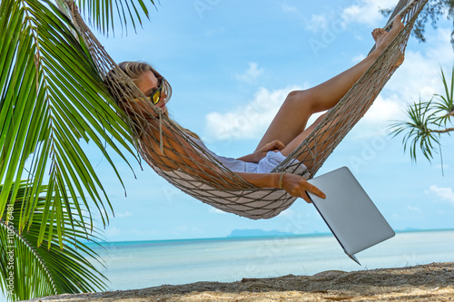 Young woman working on the computer lies in a hammock on a tropical beach