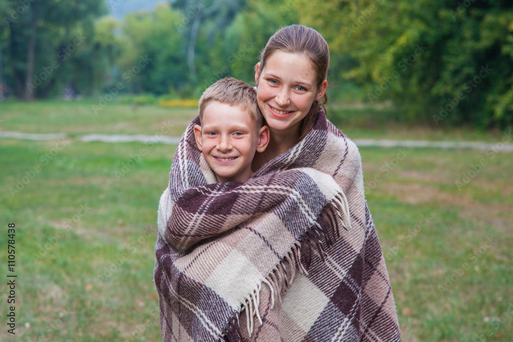Young sister and brother stand covered with plaid in park. Young sister and brother stand covered with plaid in park.