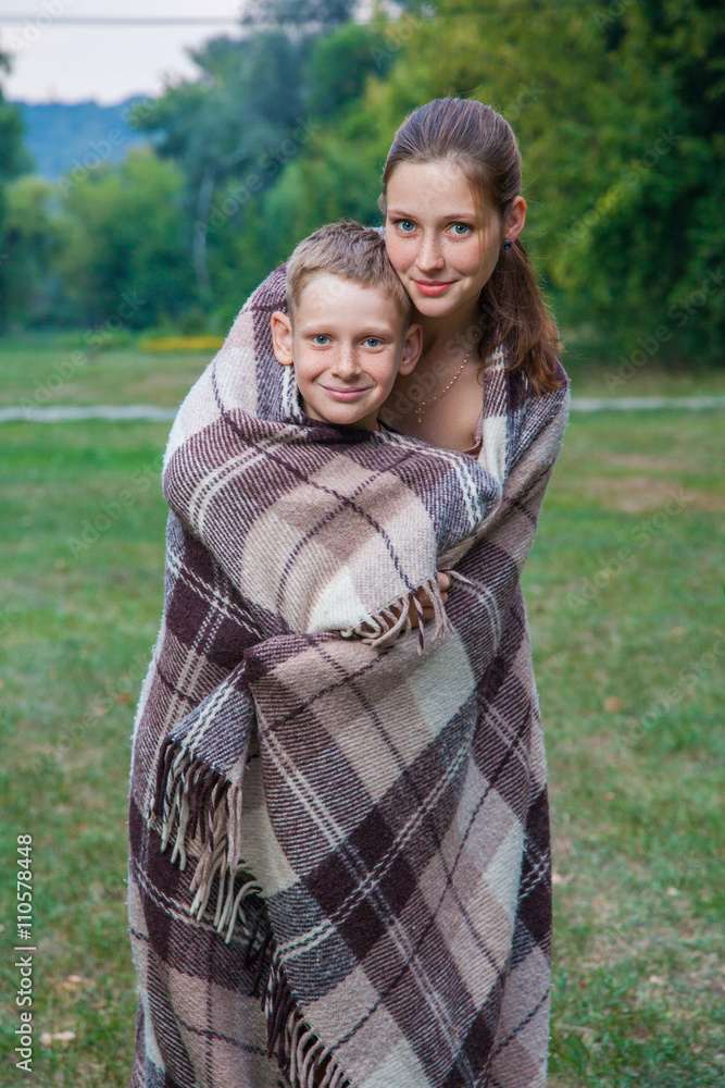 Young sister and brother stand covered with plaid in park. Young sister and brother stand covered with plaid in park.