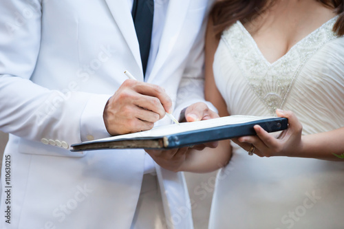 Groom and bride in white suit signing their marriage license 