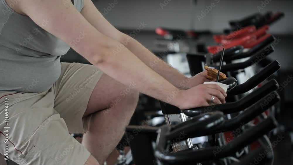 Fat guy in shorts practicing the exercise bike, holding a pizza and a glass of soda Fat guy in shorts practicing the exercise bike, holding a pizza and a glass of soda