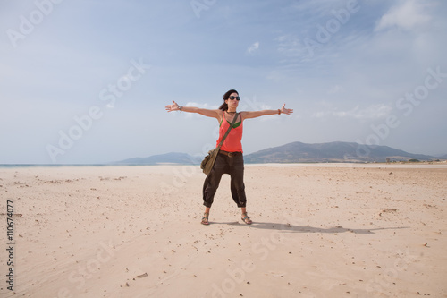 brunette summer vacation woman with sunglasses, orange shirt and brown trousers standing open arms up embracing on sand greeting wind, in Lances beach, Tarifa, Cadiz, Andalusia, Spain, Europe