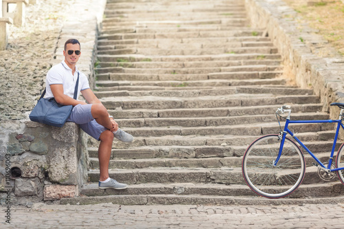 Handsome young man is sitting on the stairs of a fortress while his bicycle is beside him