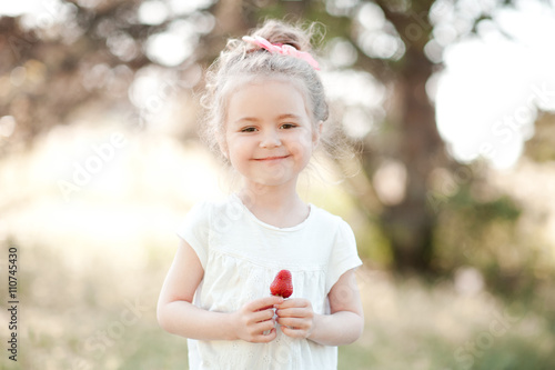 Smiling baby girl 3-4 year old holding strawberry outdoors. Looking at camera. Happiness.