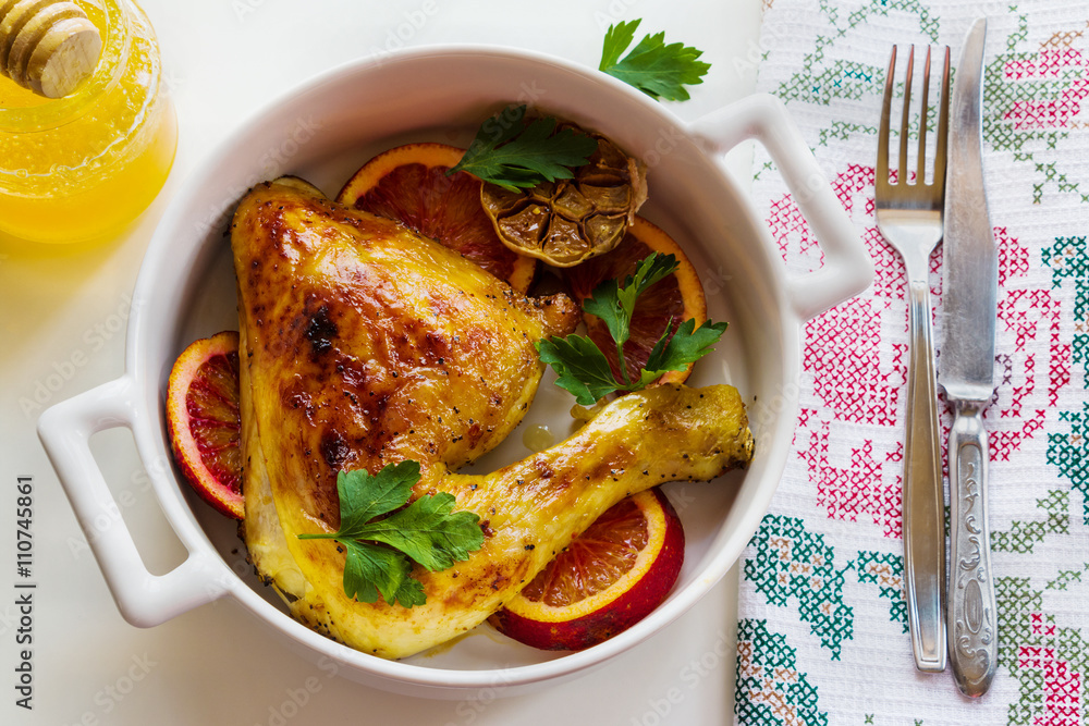 Roasted chicken legs, red oranges, garlic and parsley in white baking dish on white table. Cooked with sauce from orange, honey and olive oil. Top view. Roasted chicken legs, red oranges, garlic and parsley in white baking dish on white table. Cooked with sauce from orange, honey and olive oil. Top view.