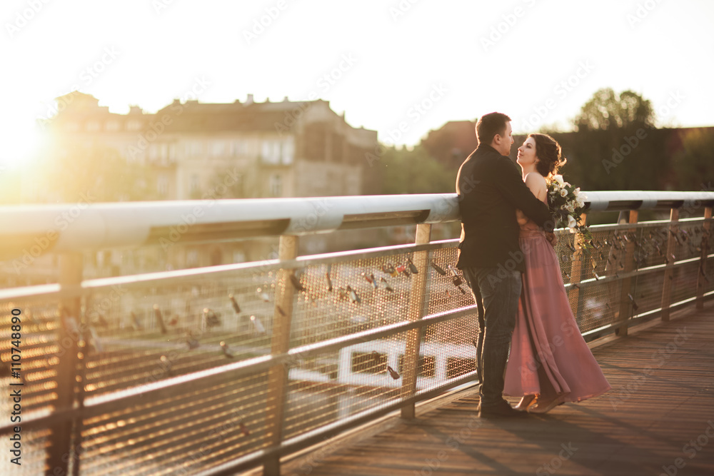 Stylish loving wedding couple, groom, bride with pink dress kissing and hugging on a bridge at sunset
