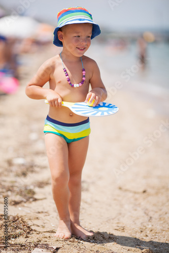 Cute boy in swim pants walking along sandy beach with badminton racket and ball