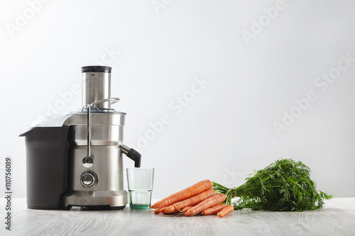 Fotografía Side view metallic professional juicer with empty glass is prepared to make tasty juice for breakfast from fresh carrots lying on wooden table
