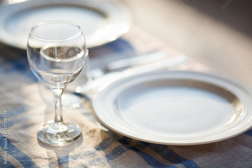 Two clean round empty plates made of porcelain near a crystal stemmed glass on a checkered tablecloth with blue lines Two clean round empty plates made of porcelain near a crystal stemmed glass on a checkered tablecloth with blue lines