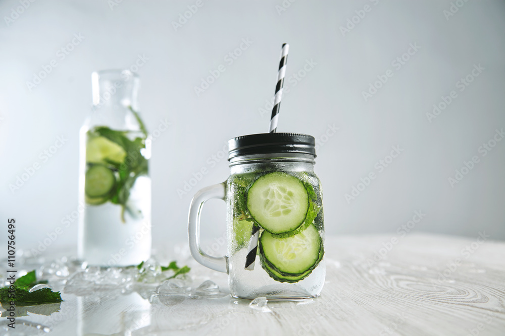 Cucumber mojito lemonade without alcohol. Fresh summer drink to cool down with mint, mineral water and ice. Rustic jar in front and bottle with beverage behind Cucumber mojito lemonade without alcohol. Fresh summer drink to cool down with mint, mineral water and ice. Rustic jar in front and bottle with beverage behind