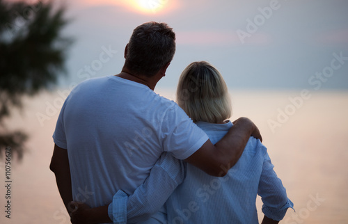 Romantic middle-aged couple standing arm in arm with their backs to the camera enjoying the sunset and a tender moment together