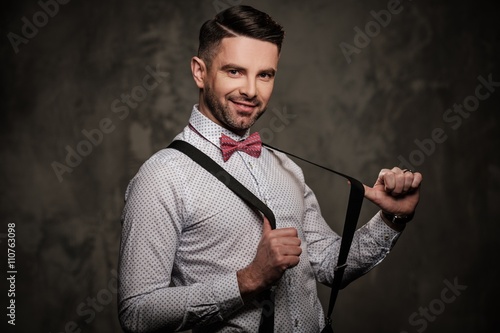 Stylish man with bow tie wearing suspenders and posing on dark background.
