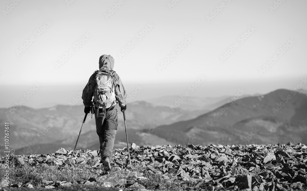 Male sportsman backpaker walking on the rocky top of the mountain with beautiful mountains on background. Man is wearing jacket and has trekking sticks and backpack on. black and white