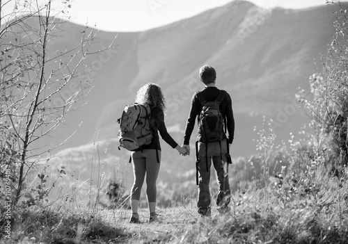 Rear view of young backpackers couple looking into distance enjoying the overlook view and holding hands on a sunny day in the mountains. Mountain on the background. black and white