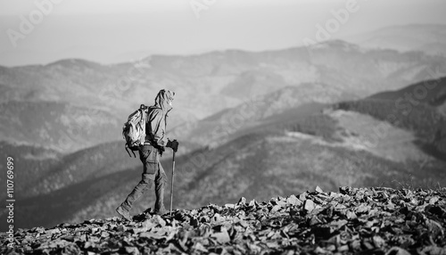 Male sportsman backpaker walking on the rocky mountain ridge with beautiful mountains on background. Man is wearing jacket and has trekking sticks and backpack on. Sunny day. black and white photo
