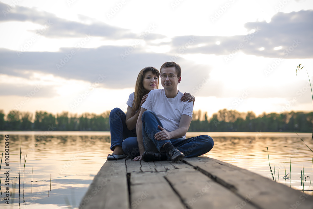 Smiling couple resting on the bridge on the shore of the lake at sunset