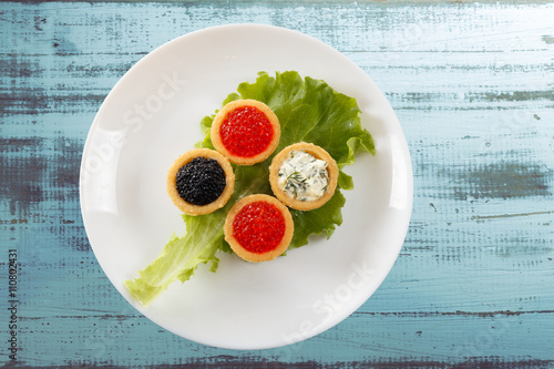 Tartlets filled with red and black caviar and cheese and dill salad on white plate against rustic wooden background