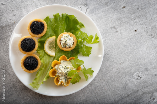 Tartlets filled with red and black caviar and cheese and dill salad on white plate against silver wooden background