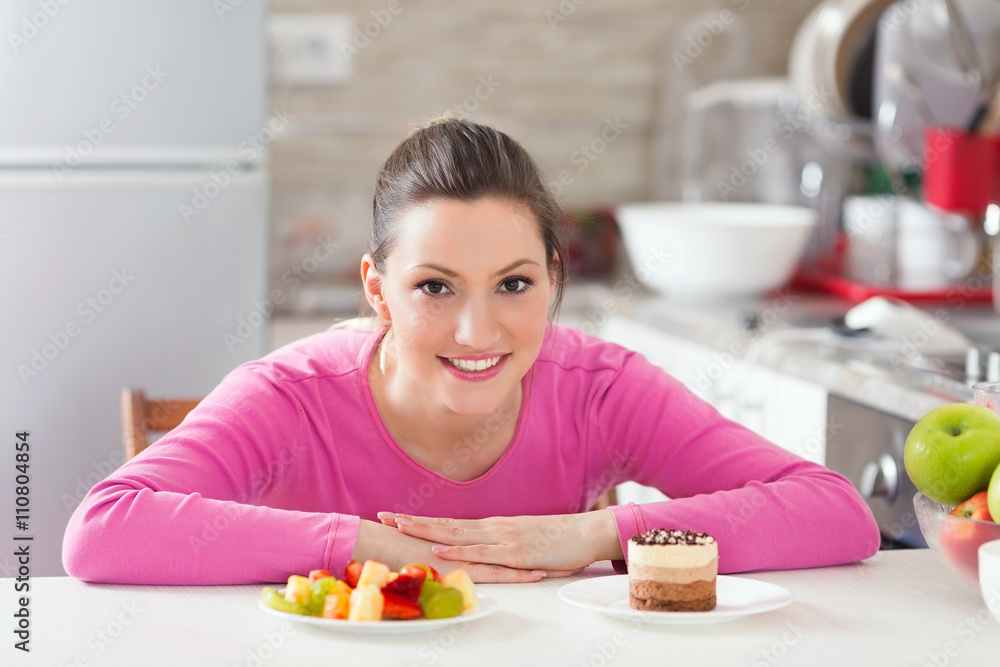 Beautiful smiling young woman is sitting at the table in the kitchen and trying to choose what to eat – healthy and fresh fruit salad or delicious chocolate cake. Beautiful smiling young woman is sitting at the table in the kitchen and trying to choose what to eat – healthy and fresh fruit salad or delicious chocolate cake.