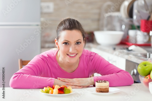 Beautiful smiling young woman is sitting at the table in the kitchen and trying to choose what to eat     healthy and fresh fruit salad or delicious chocolate cake.