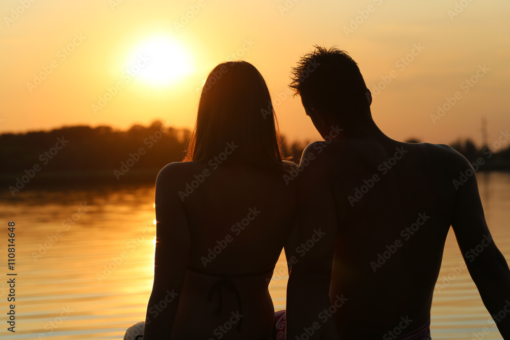 young couple sitting on lake pier, sunset silhouette
