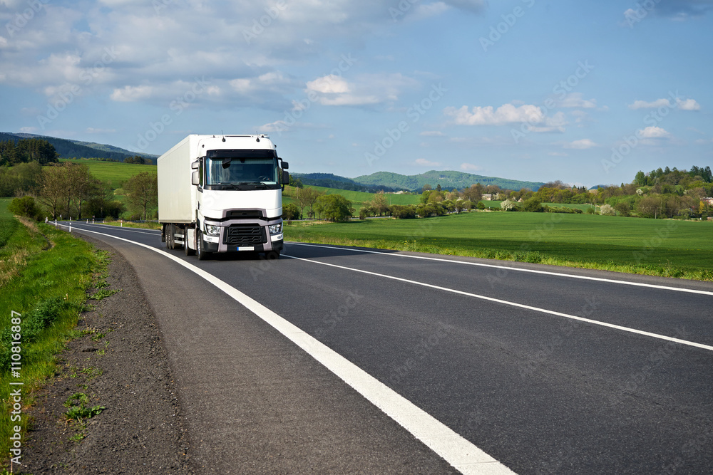 Asphalt road meander trough the green fields and white truck arriving. Villages and forested mountains in the background. Sunny spring day with blue skies and white clouds.