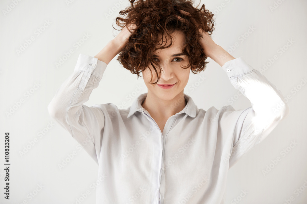 Joyful Caucasian female model shaking head with brunette curly hair wearing trendy white shirt. Pretty girl looking and smiling at the camera, having fun isolated against white studio wall background Joyful Caucasian female model shaking head with brunette curly hair wearing trendy white shirt. Pretty girl looking and smiling at the camera, having fun isolated against white studio wall background
