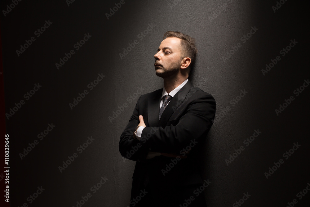 Fototapeta premium Young businessman in black suit posing with crossed or folded arms and looking away over grey background in studio.