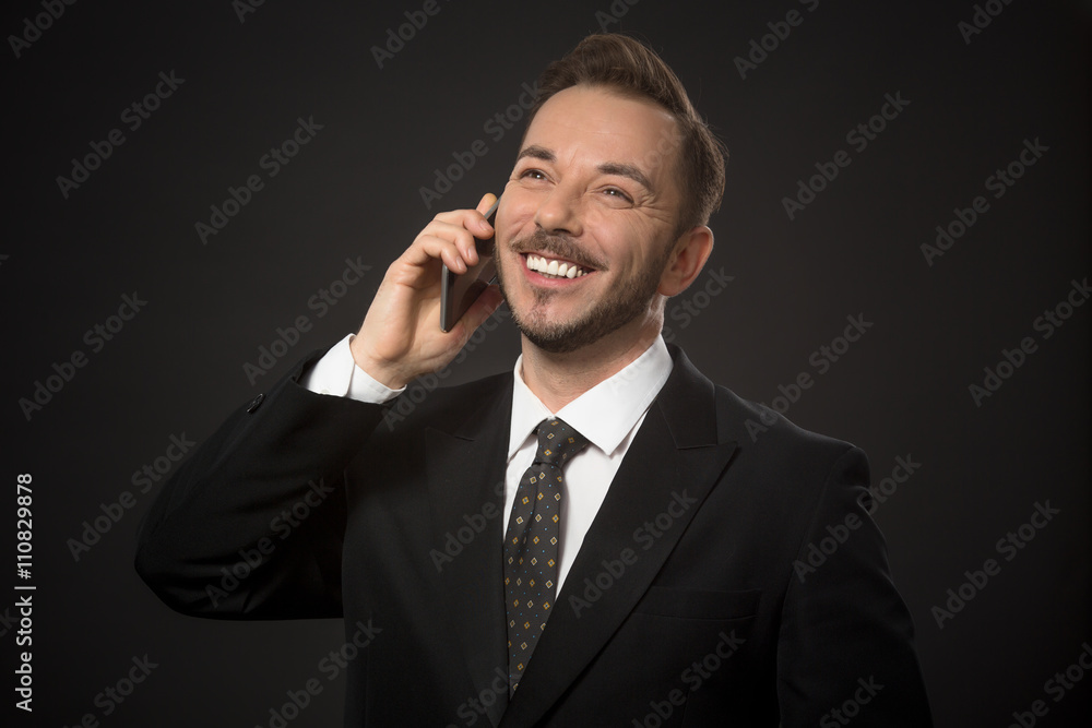 Happy businessman in black suit speaking over mobile or smart phone and toothy smiling in studio. Short-haired man speaking with his business partners. Happy businessman in black suit speaking over mobile or smart phone and toothy smiling in studio. Short-haired man speaking with his business partners.
