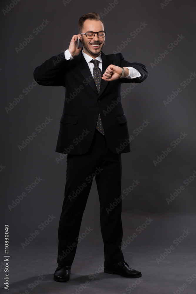 Portrat of businessman posing in full length, talking over mobile phone and looking at his watch in studio. Happy man smiling. Portrat of businessman posing in full length, talking over mobile phone and looking at his watch in studio. Happy man smiling.