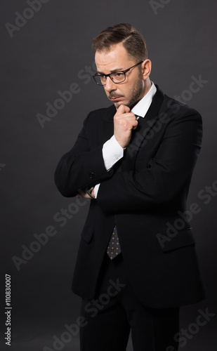 Portrait of thinkative businessman in glasses in studio. Handsome man in black suit looking down and dreaming about something.
