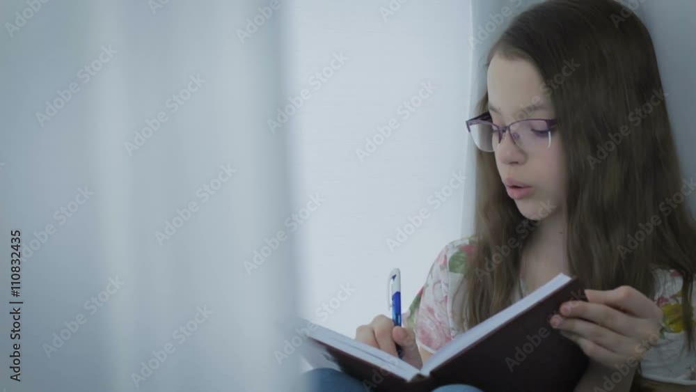 Beautiful little girl in glasses diligently does his homework at window. Beautiful little girl in glasses diligently does his homework at window.