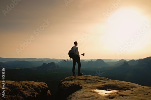 Tall ginger hair hiker in grey t-shirt and dark trekking trousers on cliff. Man with tourist poles in hand and sporty backpack above valley.