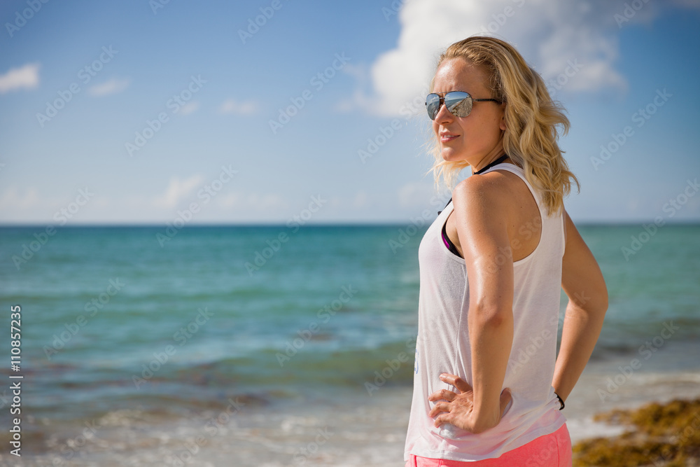 Naklejka premium Young sportive woman in sun glasses standing on the beach and looking into distance. Vacations by the sea. Summer camp. Girl relaxing after work out outdoors.