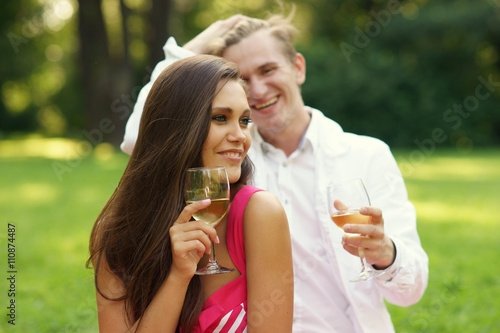 Young beautiful caucasian couple hugging and drinking wine at picnic in summer park on green grass
