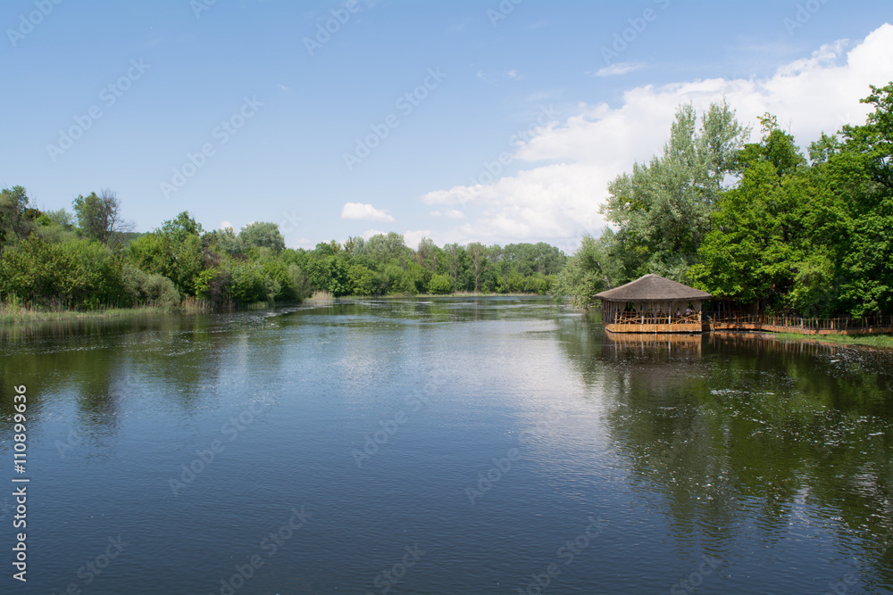 River bank: spring landscape. Blue sky, green trees and small wooden summerhouse on the water