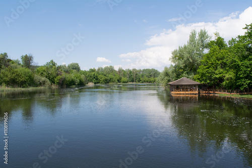 River bank: spring landscape. Blue sky, green trees and small wooden summerhouse on the water