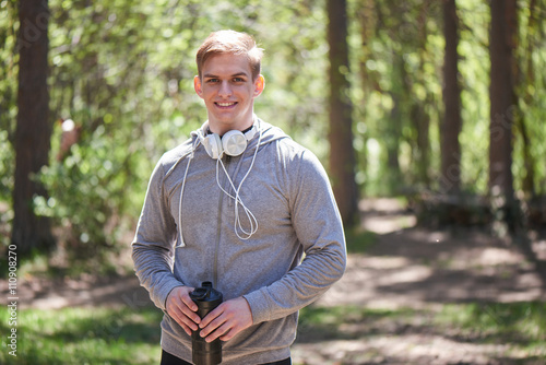 Fototapeta Naklejka Na Ścianę i Meble -  Portrait of a smiling athlete with headphones around his neck holding the shaker in hand on a forest background