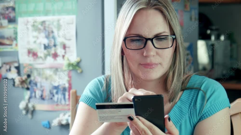 Beautiful blonde adult caucasian woman sitting at home in kitchen with stylish eyeglasses with smartphone and credit card shopping online in internet using app on cell phone. She happy to buy purchase Beautiful blonde adult caucasian woman sitting at home in kitchen with stylish eyeglasses with smartphone and credit card shopping online in internet using app on cell phone. She happy to buy purchase