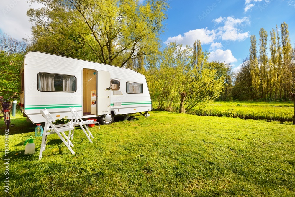 White caravan trailer on a green lawn in a camping site. Sunny day. Spring landscape. Europe. Lifestyle, travel, ecotourism, road trip, journey, vacations, recreation, transportation, RV, motorhome - obrazy, fototapety, plakaty 
