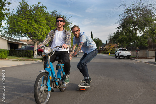 Male skateboarder skitching on friends moped on suburban road