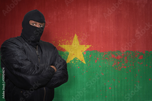 dangerous man in a mask standing near a wall with painted national flag of burkina faso