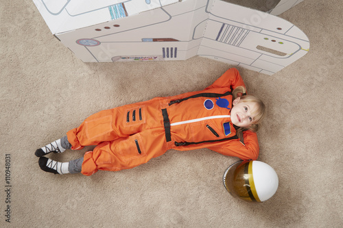 Young girl playing, wearing astronaut outfit, lying next to cardboard spaceship, elevated view photo