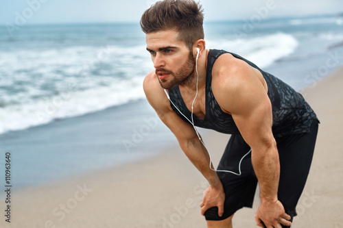 Portrait Of Athletic Man With Fit Muscular Body Resting After Jogging On Beach. Tired Exhausted Male Runner Taking A Break, Breathing After Running Workout Outdoor Near Ocean. Sports, Fitness Concept
