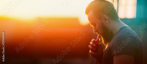 young man with vaporiser  tightening pairs on sunset sky background, blurred background, banner for web site