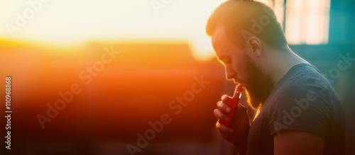 Obraz na plátně young man with vaporiser  tightening pairs on sunset sky background, blurred bac