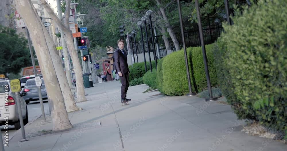 Young man in business suit rides towards from the camera on a skateboard against a city background with trees and bushes in Downtown Los Angeles. Slow motion recorded at 60fps. Young man in business suit rides towards from the camera on a skateboard against a city background with trees and bushes in Downtown Los Angeles. Slow motion recorded at 60fps.