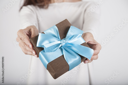 brown gift box with blue ribbon in woman blue jeans cream jersey hands over white background
